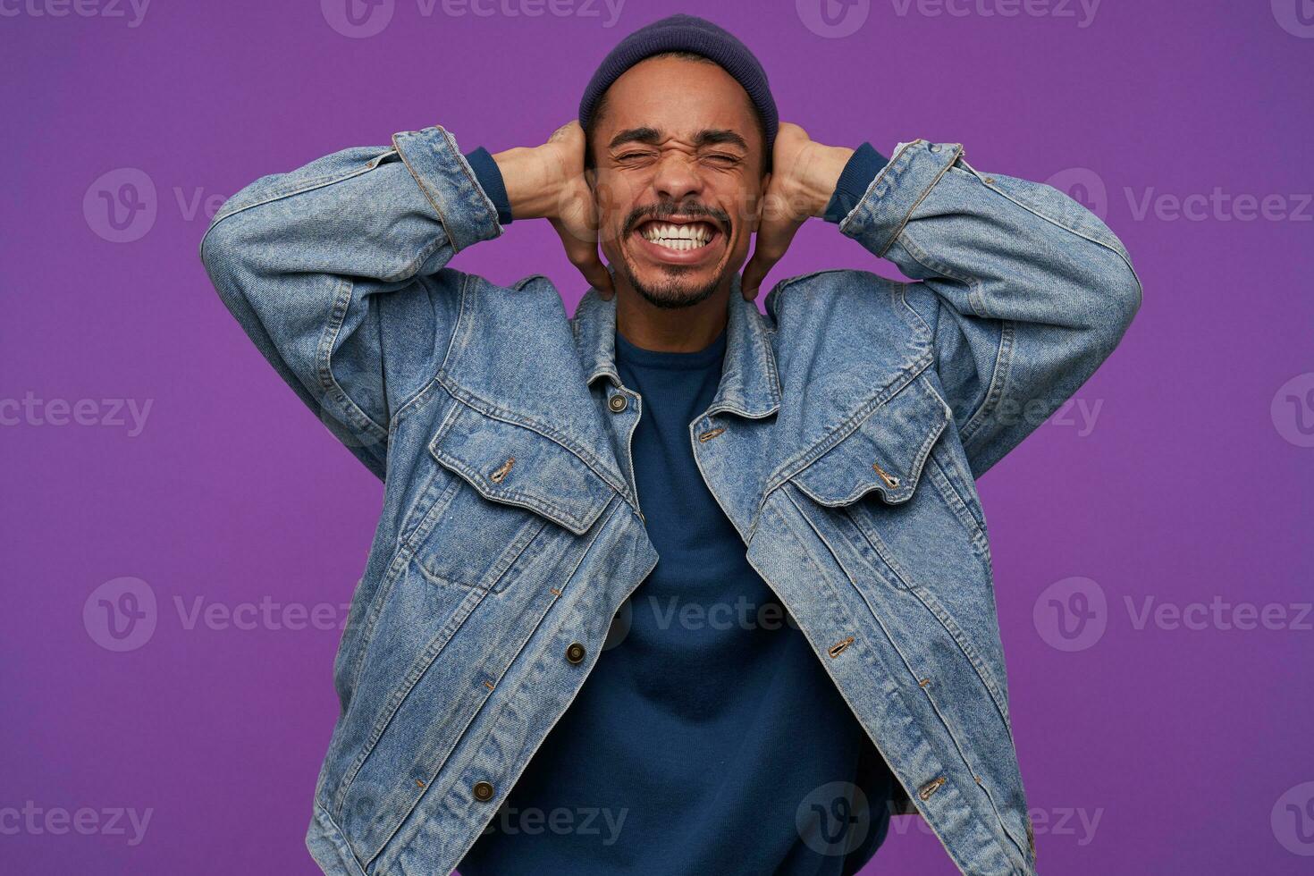 Studio shot of young stressed dark skinned bearded guy frowning his face with closed eyes while covering ears with raised palms, avoiding loud sounds, isolated over purple background photo