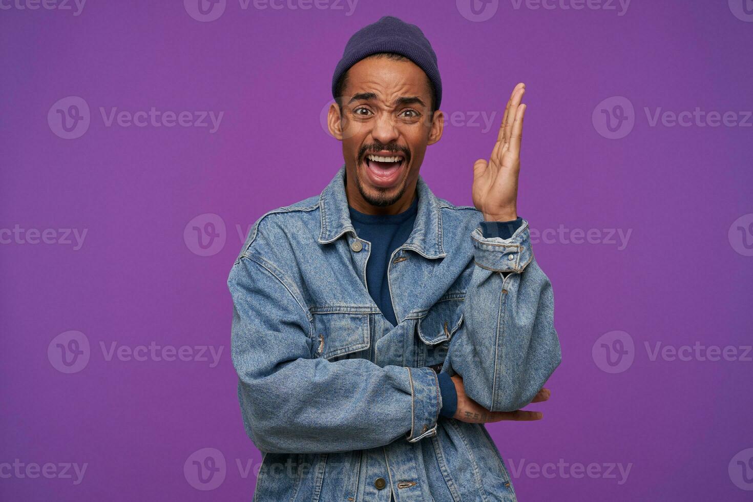 Excited young bearded brunette guy with dark skin raising hand while looking emotionally at camera with wide mouth opened and frowning his face, isolated over purple background photo