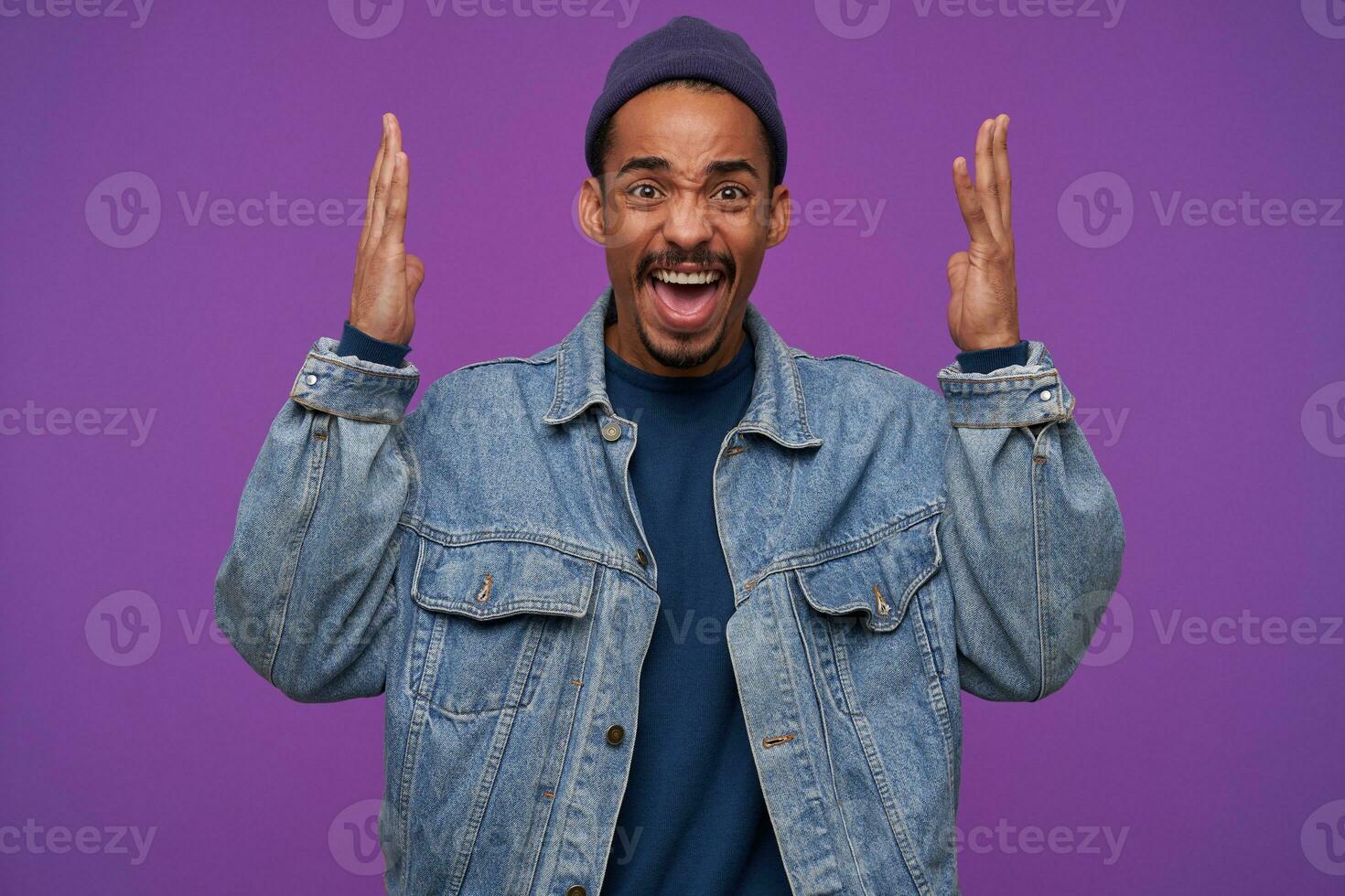 Portrait of young pretty dark skinned bearded guy raising palms emotionally and frowning face while shouting angrily, dressed in casual clothes while posing over purple background photo