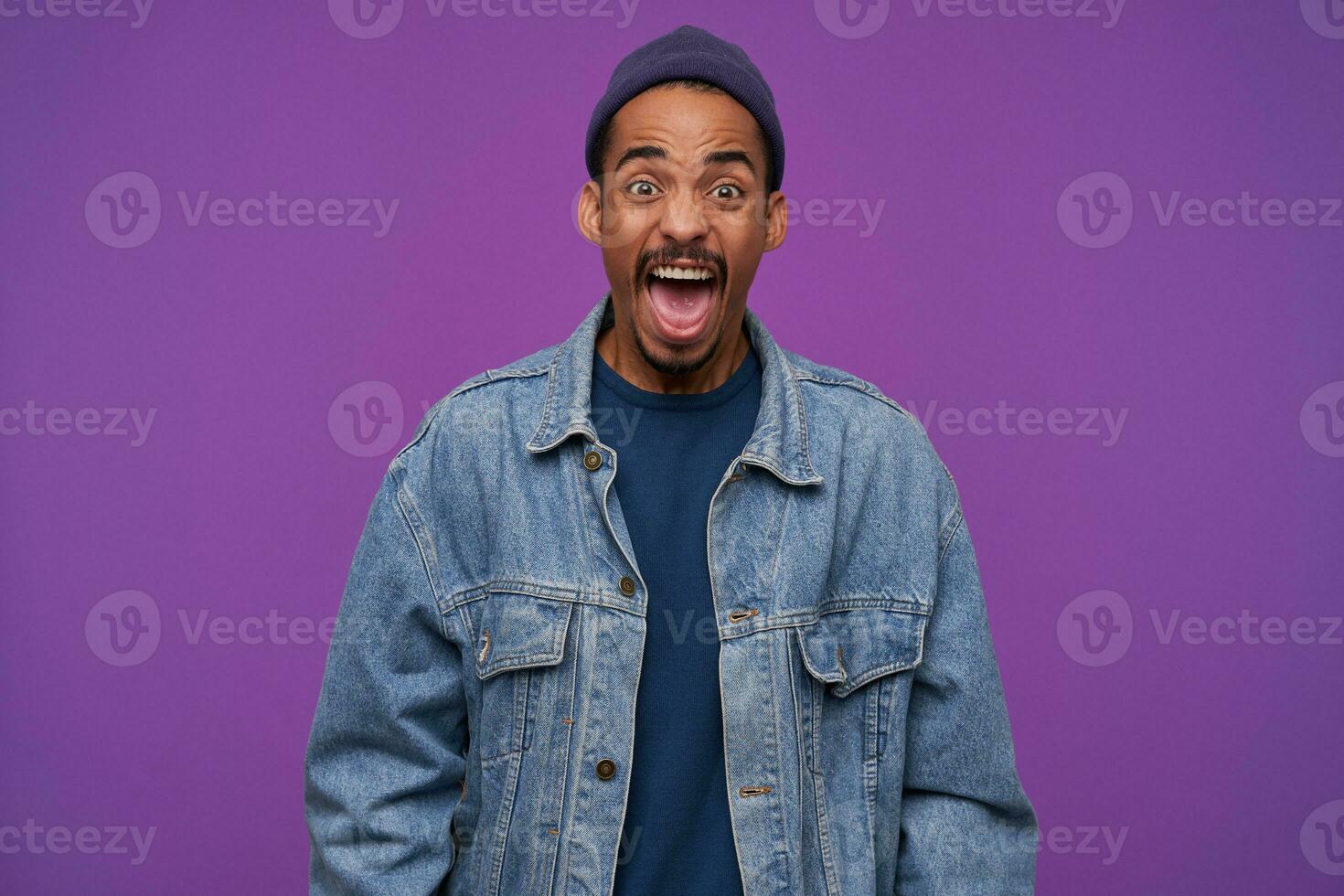 Furious young brown-eyed bearded man with dark skin shouting angrily and wrinkling forehead while looking heatedly at camera, wearing blue cap, pullover and jeans coat over purple background photo