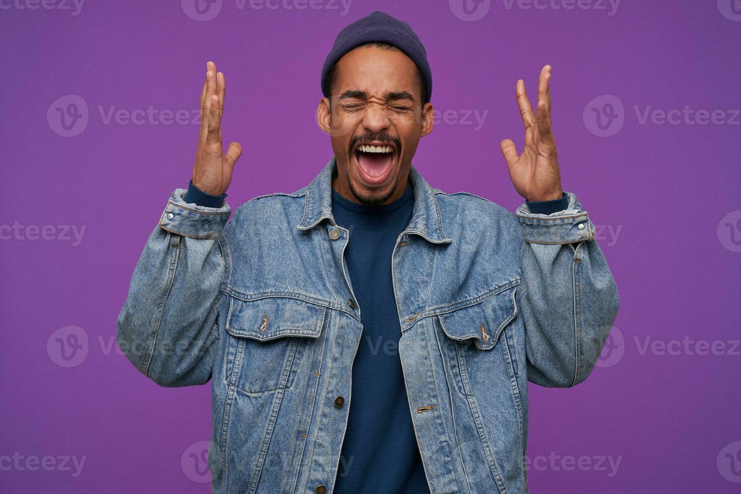 Studio photo of young stressed dark skinned bearded brunette man keeping his eyes closed while screaming furiously with raised hands, posing over purple background