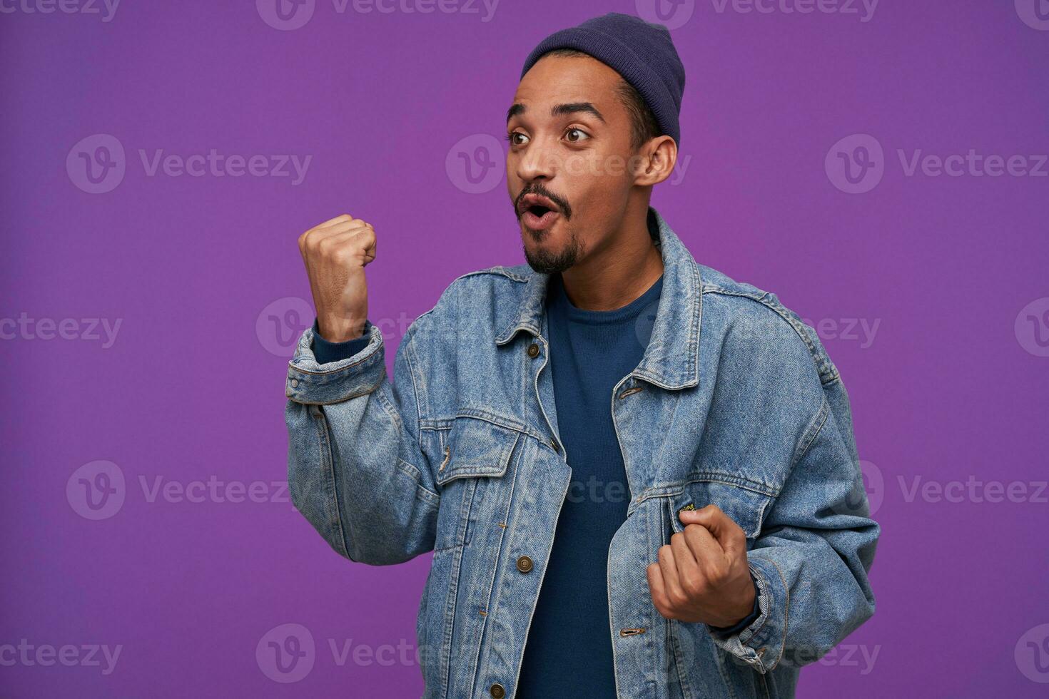 Excited young pretty dark haired bearded male showing fist while looking aside with agitated face, wearing blue cap, pullover and jeans coat while posing over purple background photo