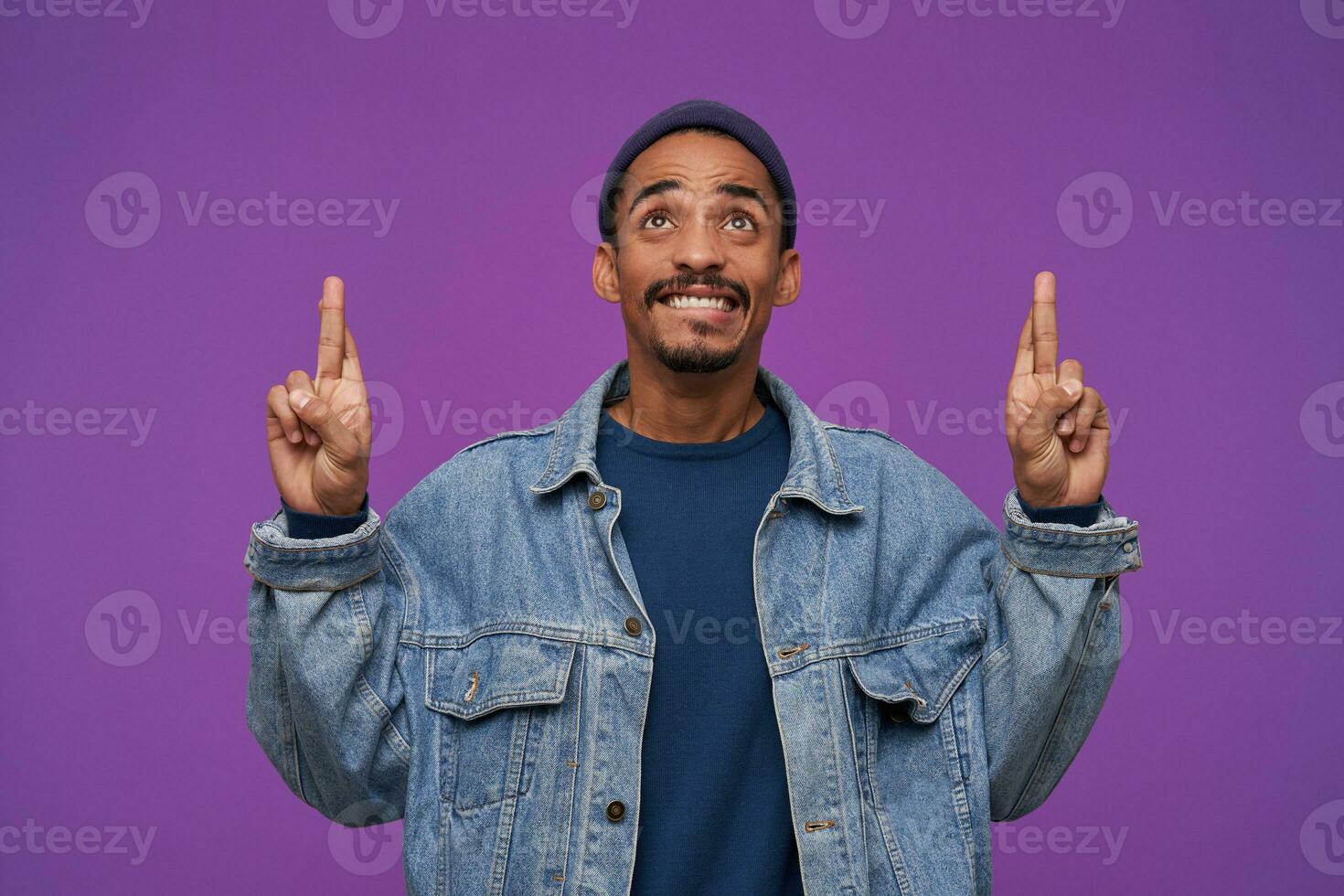 Handsome young dark skinned bearded brunette man looking upwards with big hope and biting underlip, keeping forefingers raised while posing over purple background photo