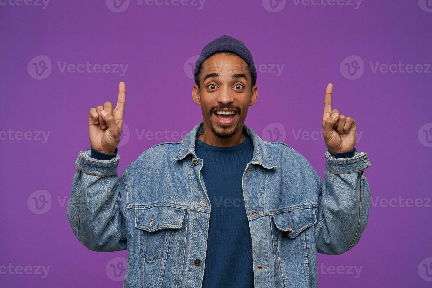 Joyful young handsome dark skinned brunette male with beard showing upwards while standing over purple background, rounding his brown eyes while looking happily at camera photo