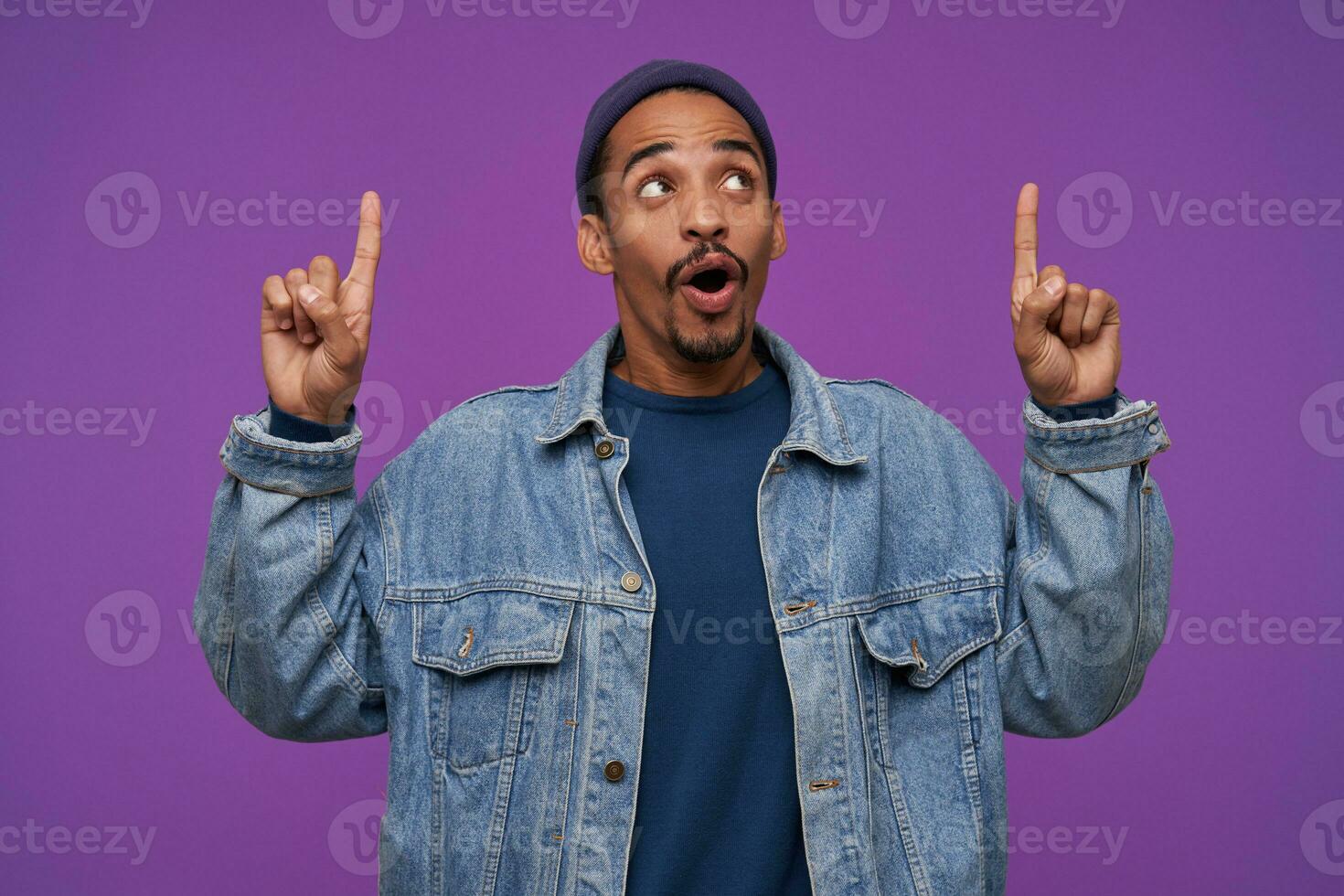 Studio photo of attractive young dark skinned brunette male with beard keeping his forefingers raised while looking upwards with excited face, isolated over purple background