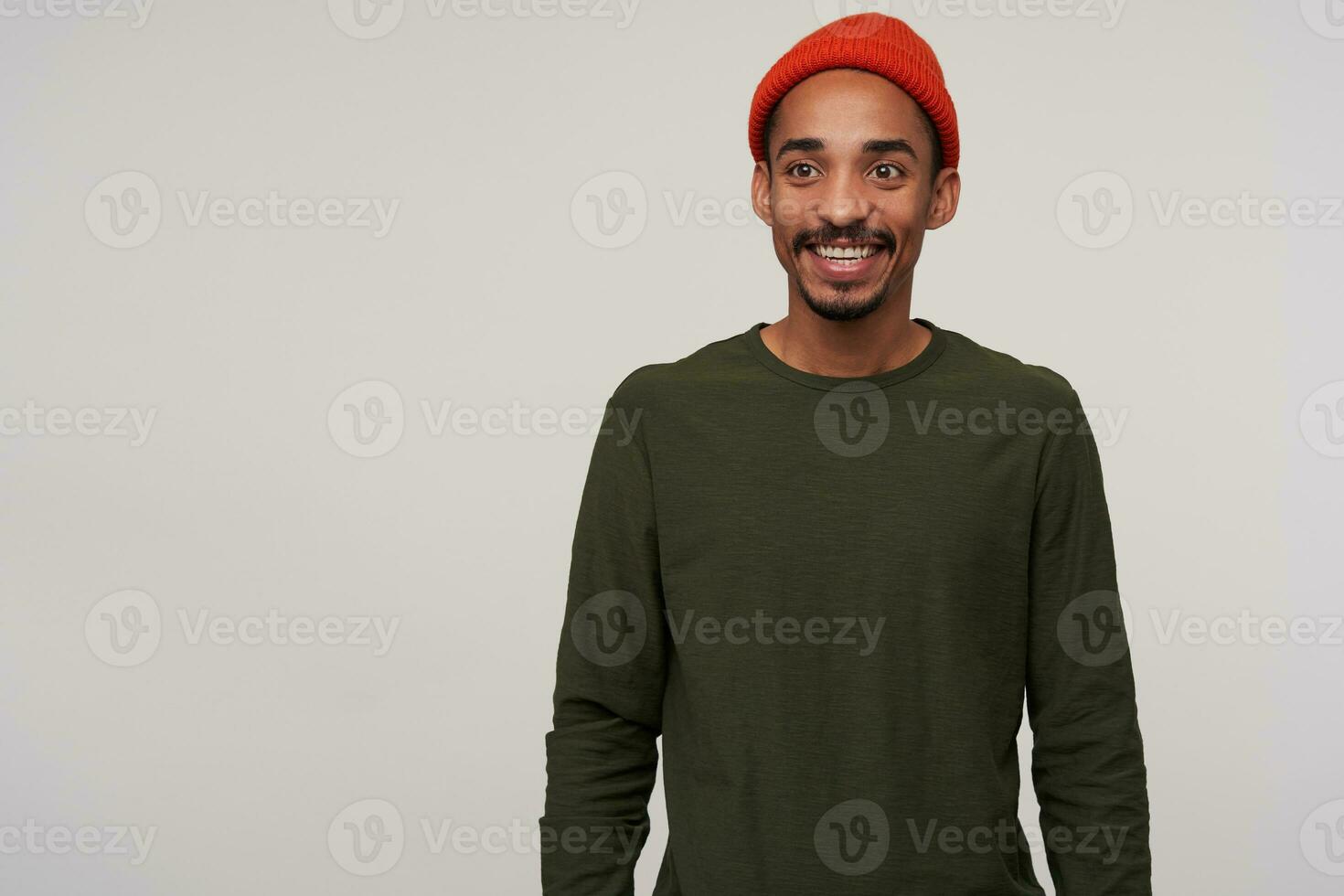 Positive young lovely bearded dark skinned brunette male in red hat and casual clothes looking aside with charming smile while standing over white background with hands down photo