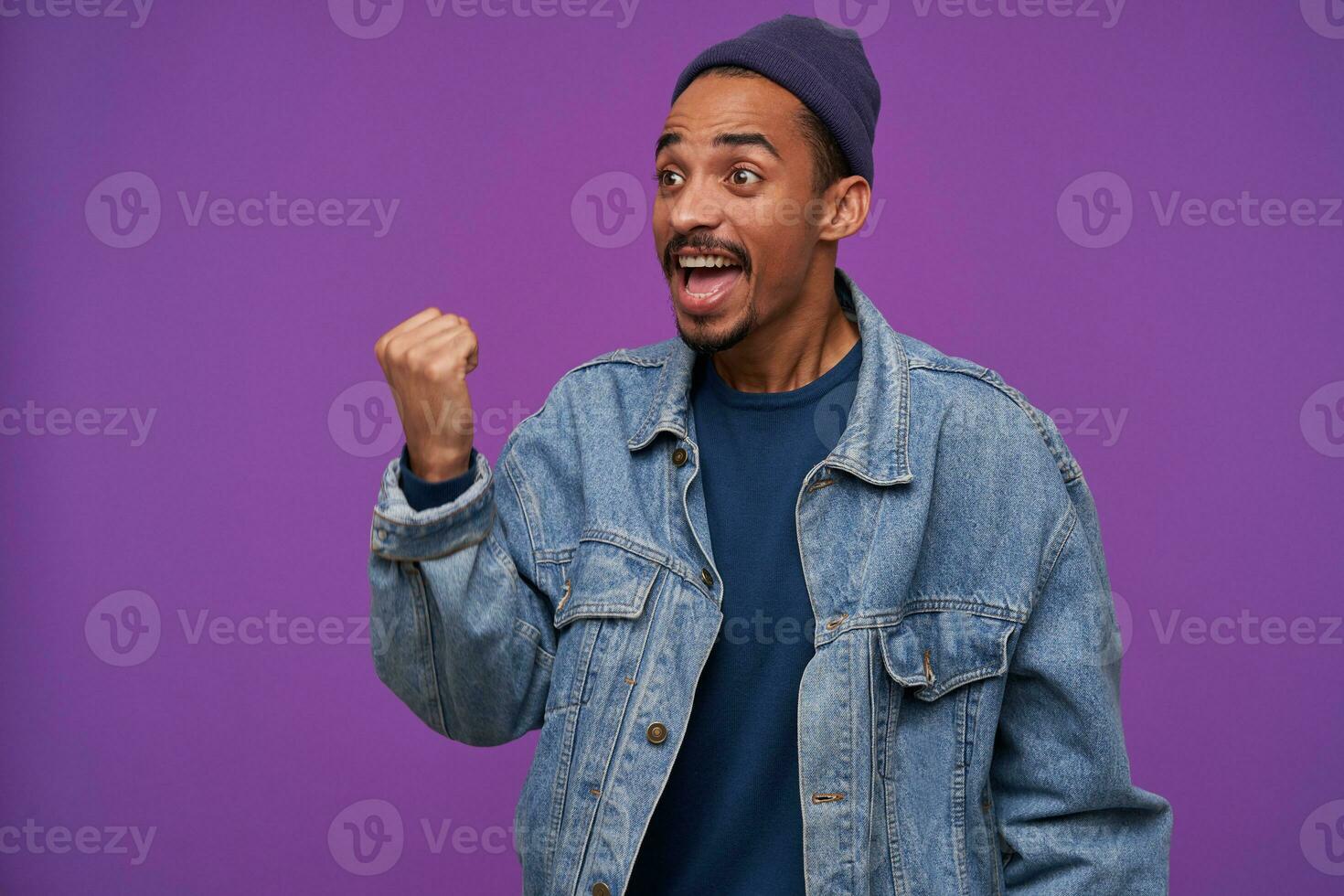 Overjoyed young lovely dark skinned bearded brunette man screaming happily and raising up his fist while looking emotionally aside, isolated over purple background photo