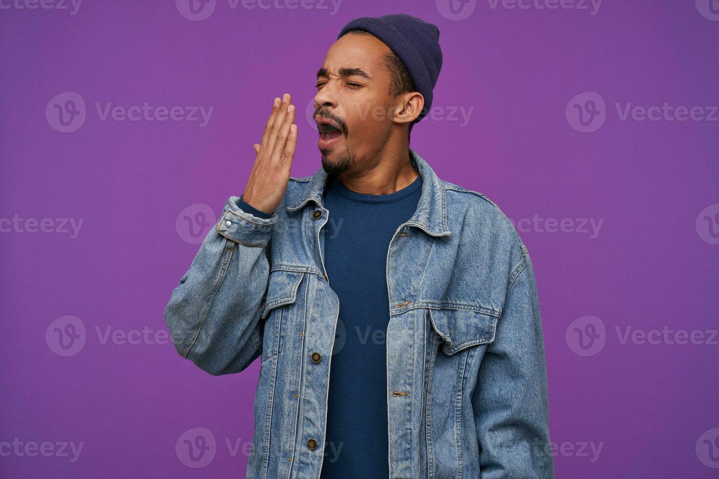 Tired young pretty dark skinned bearded brunette guy covering his mouth with raised palm while yawning with closed eyes, dressed in clothes while posing over purple background photo