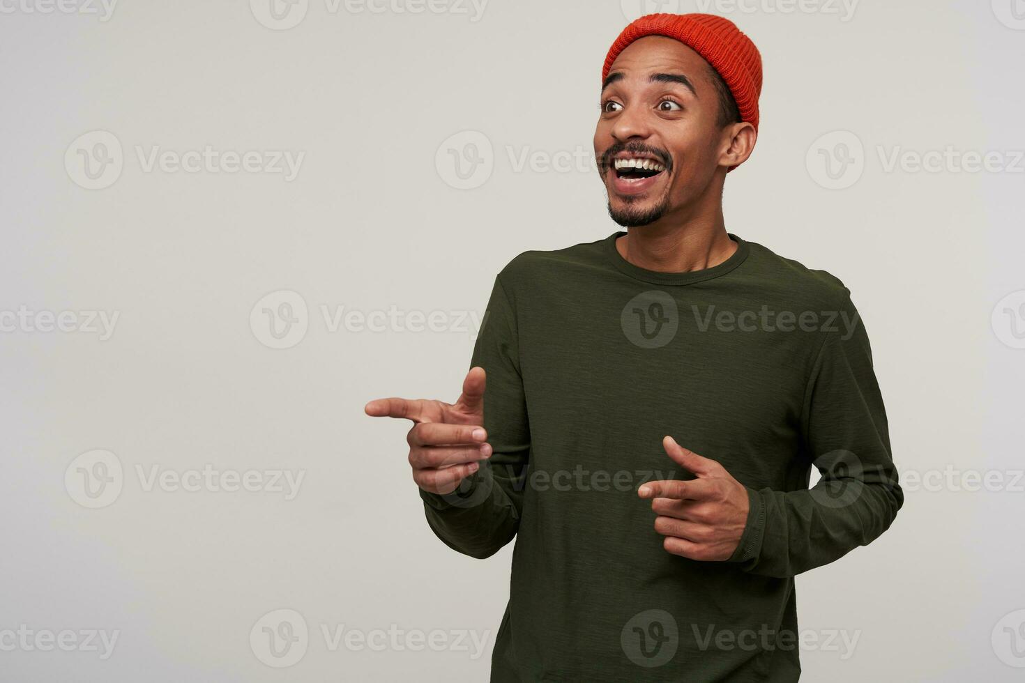 Indoor shot of young pretty joyful dark haired bearded man with dark skin showing happily aside with raised forefingers and smiling widely, standing against white background photo