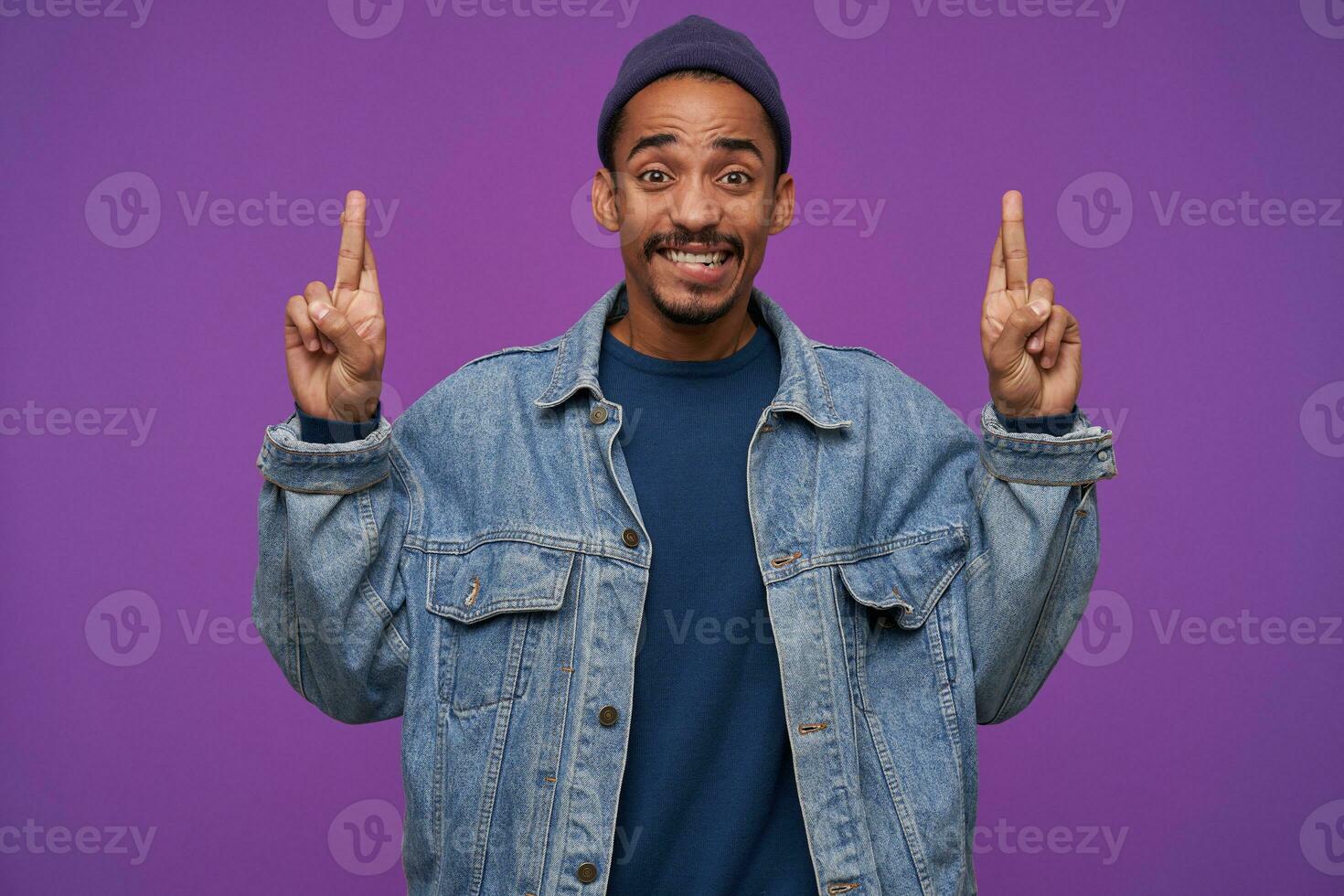 Portrait of young brown-eyed dark skinned brunette guy showing upwards with crossed fingers and looking worringly at camera, dressed in casual wear while posing over purple background photo