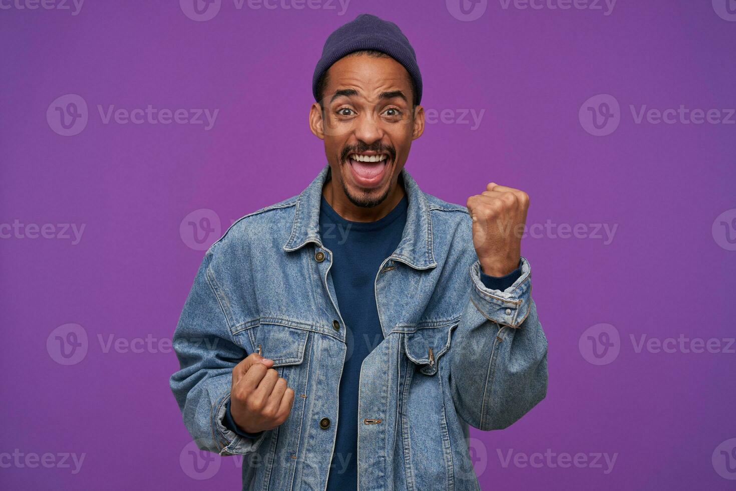 Portrait of young energized pretty dark skinned bearded male raising happily fists and screaming cheerfully with wide mouth opened, isolated over purple background photo