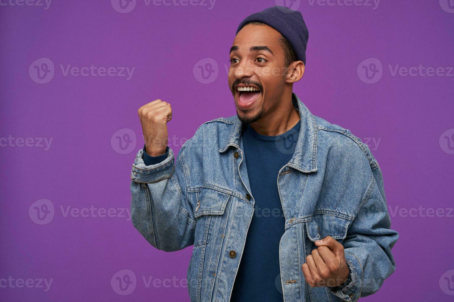 Agitated young attractive dark skinned bearded guy in blue cap and jeans coat looking aside with excited face and raising emotionally his fist, standing over purple background photo