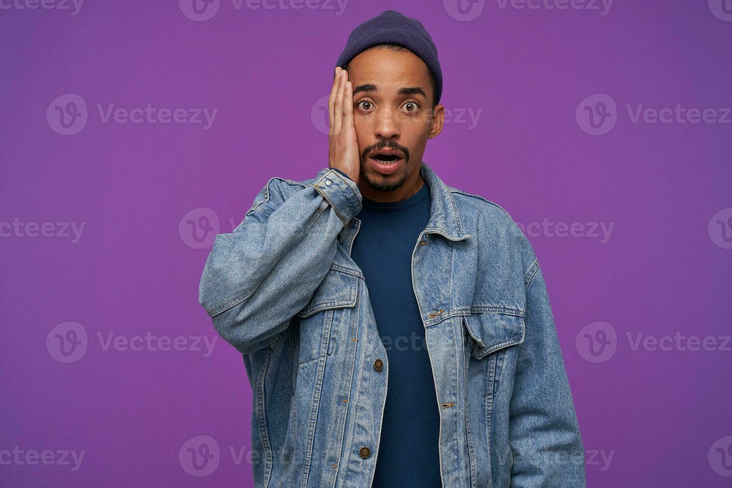 Shocked young brown-eyed dark skinned bearded man holding palm on his cheek while looking amazedly to camera with opened mouth, isolated over purple background photo