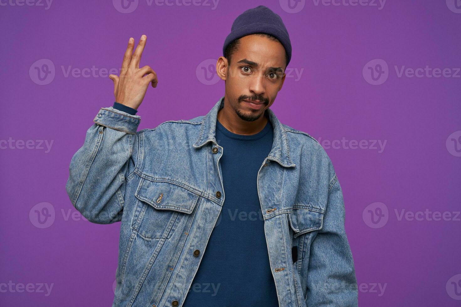 Puzzled young brown-eyed bearded hansome male with dark skin looking confusedly at camera with folded lips, posing over purple background with raised hand photo