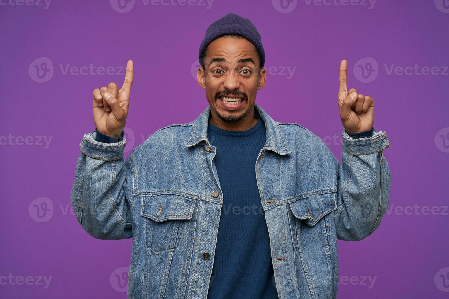 Displeased young brown-eyed bearded brunette man in casual clothes showing upwards with forefingers while looking at camera with pout, standing against purple background photo