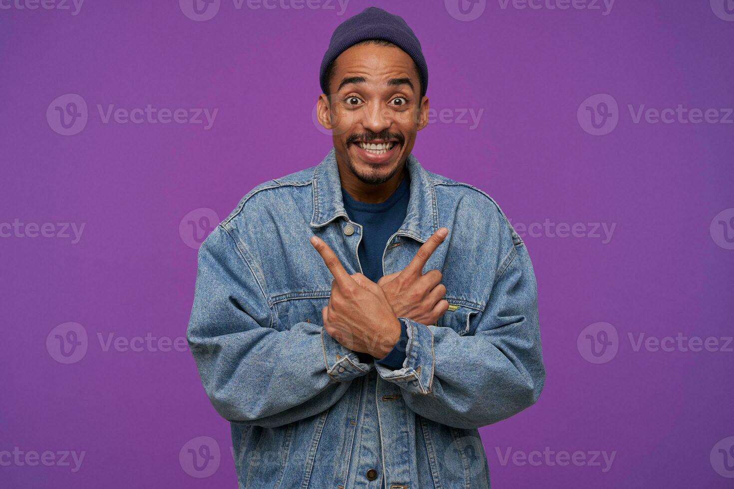 Overjoyed young brown-eyed dark skinned bearded male looking amazedly at camera with wide smile, showing in different sides with raised index fingers, isolated over purple background photo