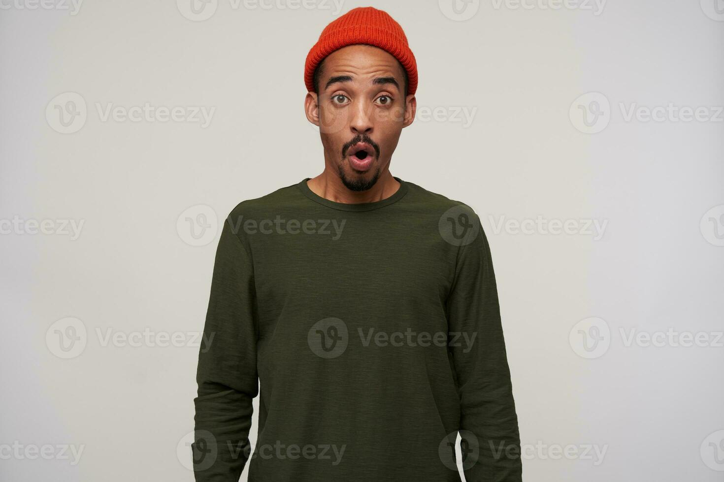 Overwhelmed young attractive bearded brunette male with dark skin looking amazedly at camera with round eyes, wearing red hat and khaki sweater over white background photo