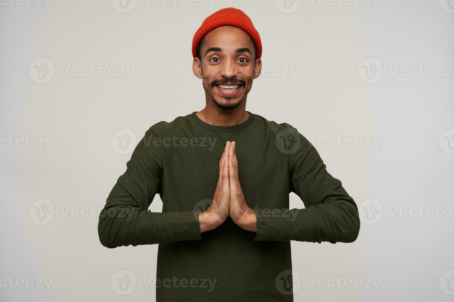 Positive young dark haired brown-eyed dark skinned man with beard folding raised palms together and looking happily at camera with wide smile, standing against white background photo