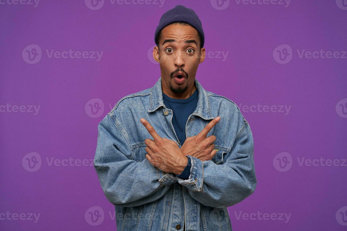 Open-eyed young pretty dark skinned bearded guy crossing hands on his chest and raising surprisedly eyebrows while looking amazedly at camera, isolated over purple background photo