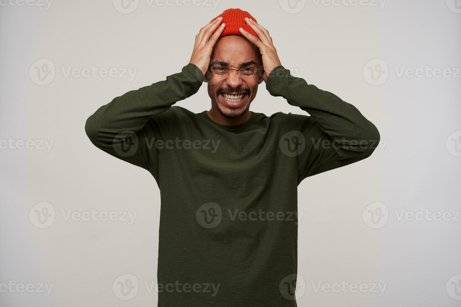 Confused young brown-eyed bearded dark skinned brunette guy frowning his face and holding head with raised hands while looking at camera, isolated over white background photo