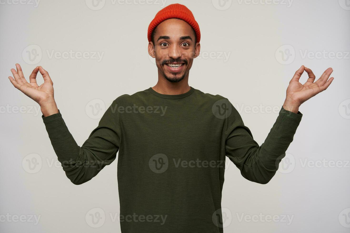 Indoor shot of cheerful attractive young brown-eyed dark skinned man with beard smiling widely at camera and raising hands with mudra sign, isolated over white background photo