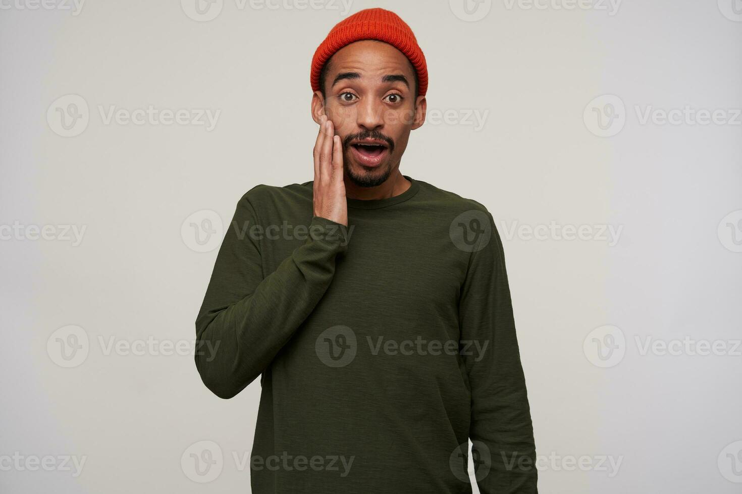 Open-eyed young dark haired dark skinned male with beard holding raised palm on his cheek while looking surprisedly to camera, standing against white background photo