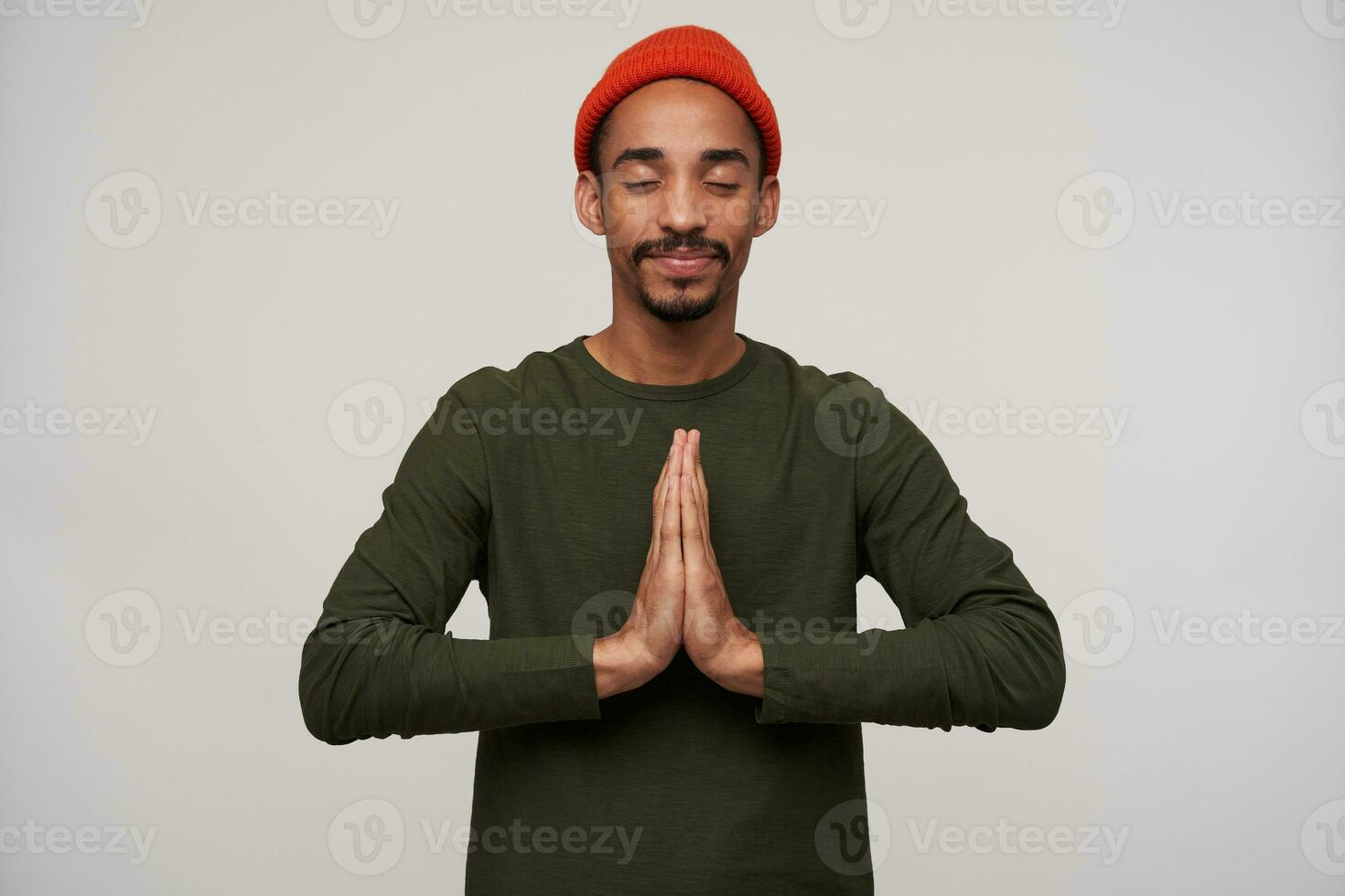 Pleasant looking young bearded dark skinned brunette guy raising hands in praying gesture while posing over white background, keeping eyes closed and smiling slightly photo