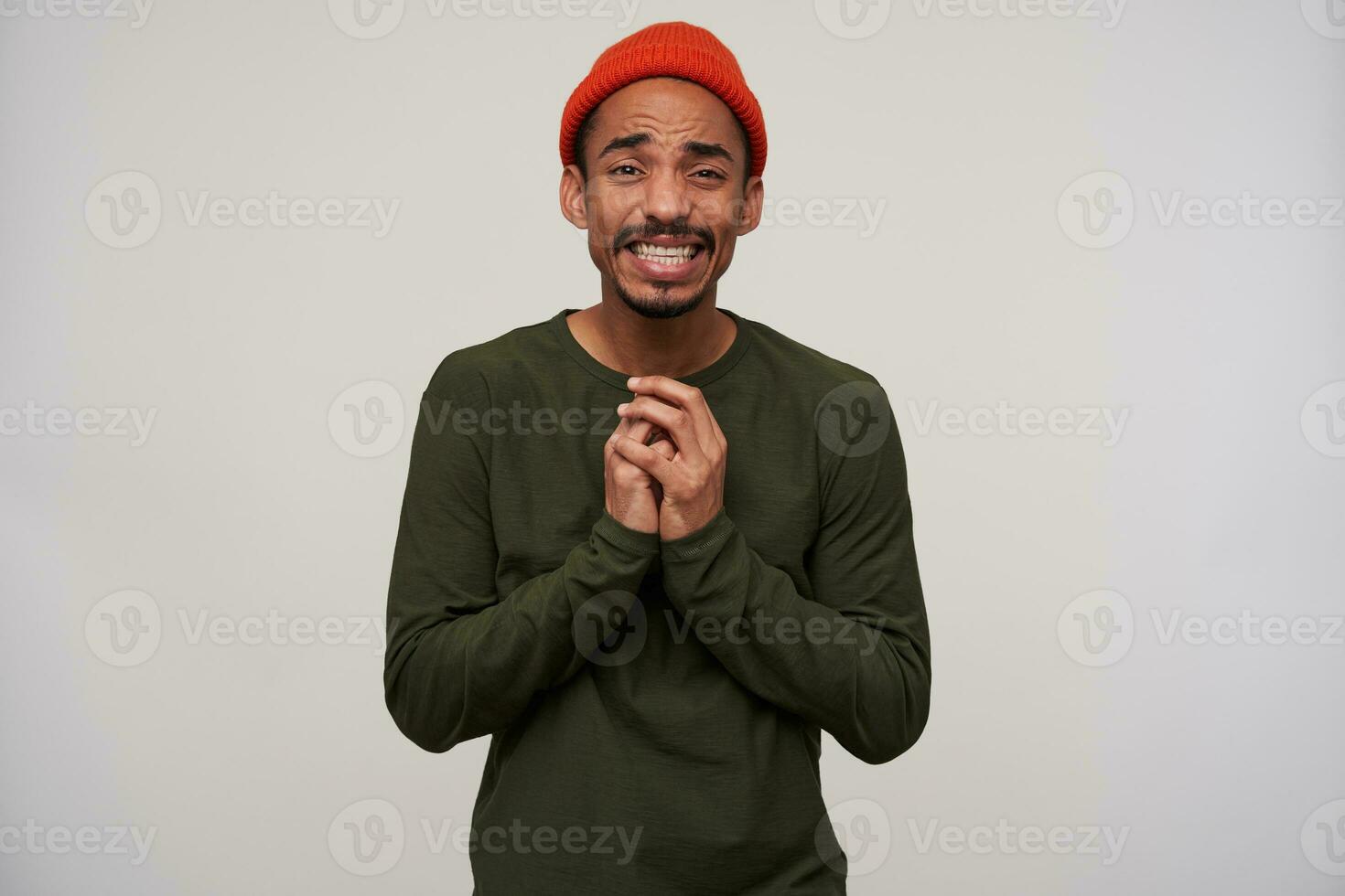 Portrait of young pretty bearded dark skinned brunette male folding hands in begging gesture and looking pityingly at camera, wearing casual clothes while standing over white background photo