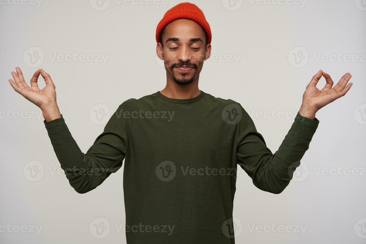 Good looking young dark skinned brunette male with beard meditating with closed eyes and folding fingers in mudra sign, wearing red hat and khaki pullover over white background photo