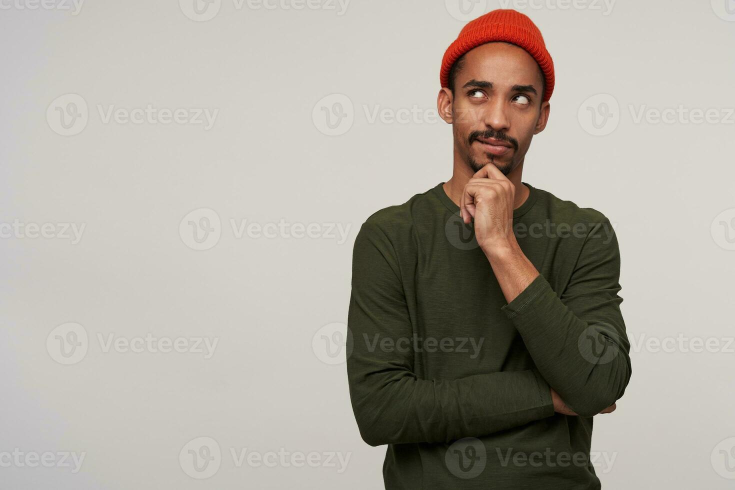 Studio photo of puzlled young pretty dark skinned man with beard holding raised hand on his chin and looking thoughtfully upwards, isolated over white background