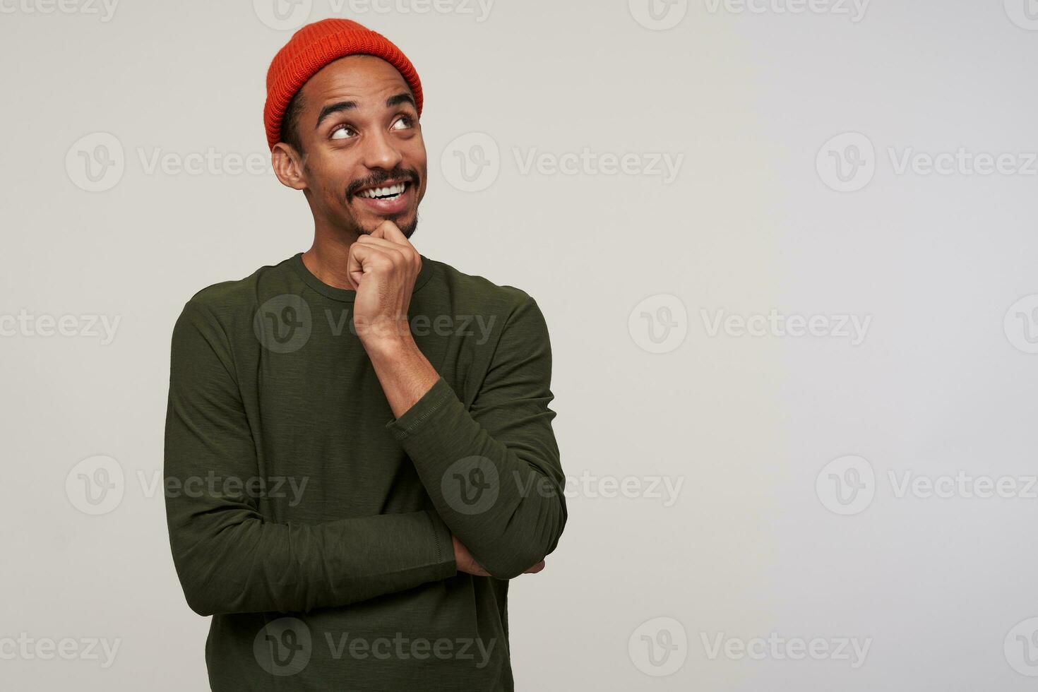 Cheerful young lovely dark haired bearded male with dark skin keeping hand on his chin and smiling pleasantly, dressed in red hat and khaki sweater while standing over white background photo