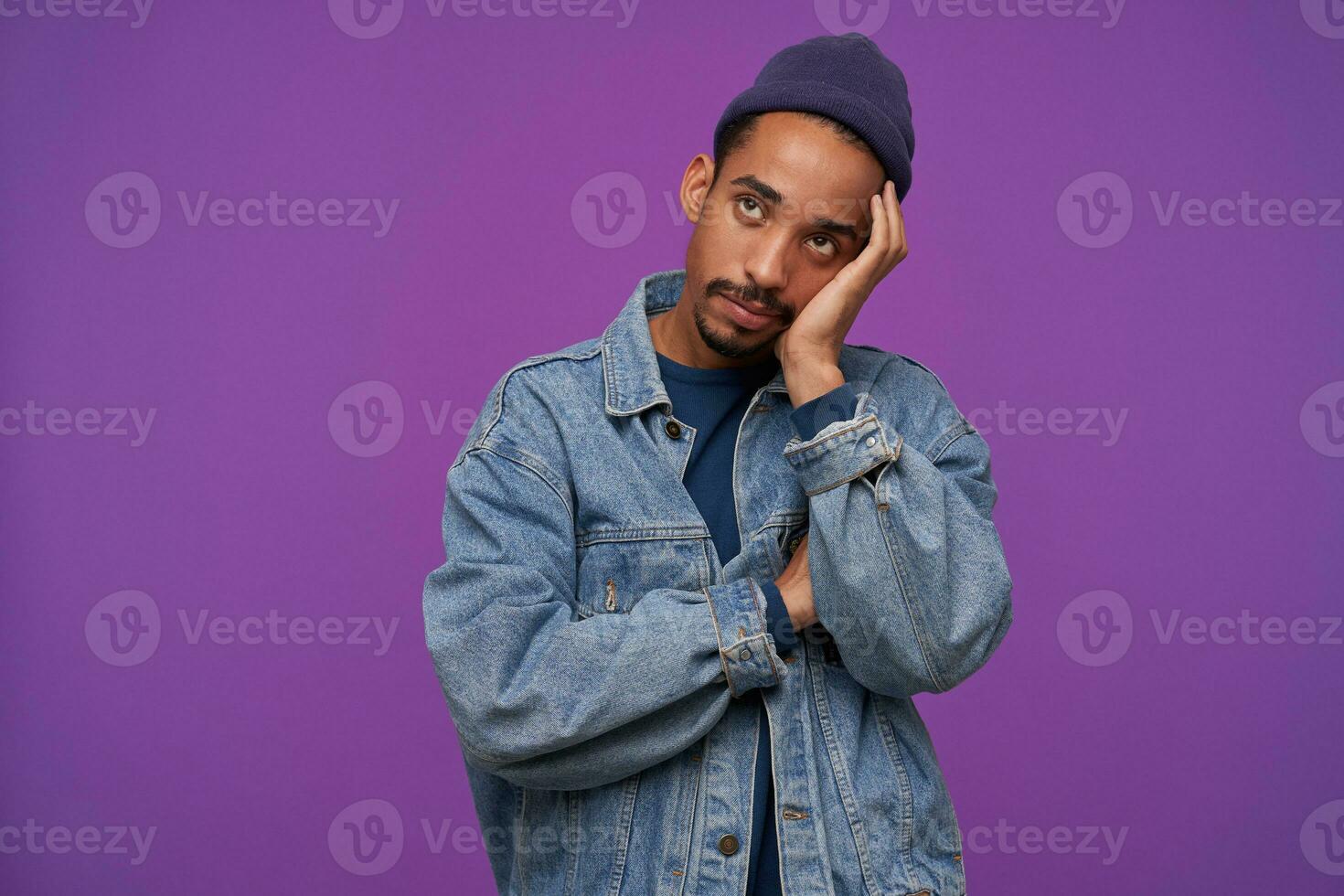 Bored young handsome bearded brunette guy with dark skin leaning his head on raised palm and looking upwards with folded lips, wearing blue cap, pullover and jeans coat over purple background photo