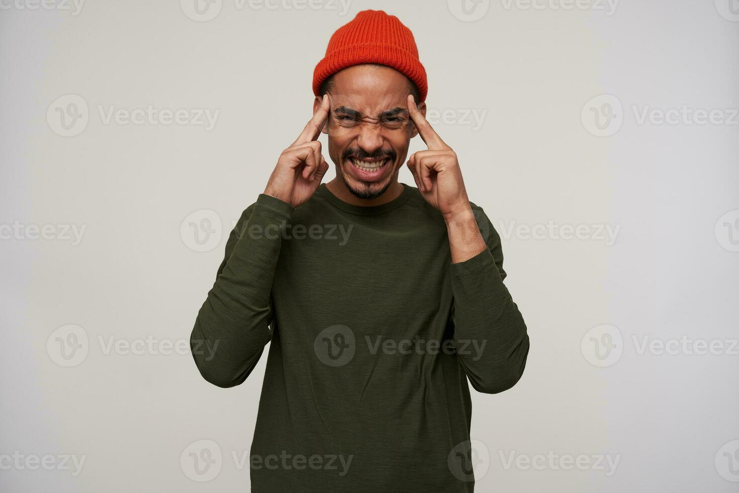 Dissatisfied young bearded dark skinned brunette guy looking painfully at camera and showing his perfect white teeth, holding forefingers on temples, standing against white background photo