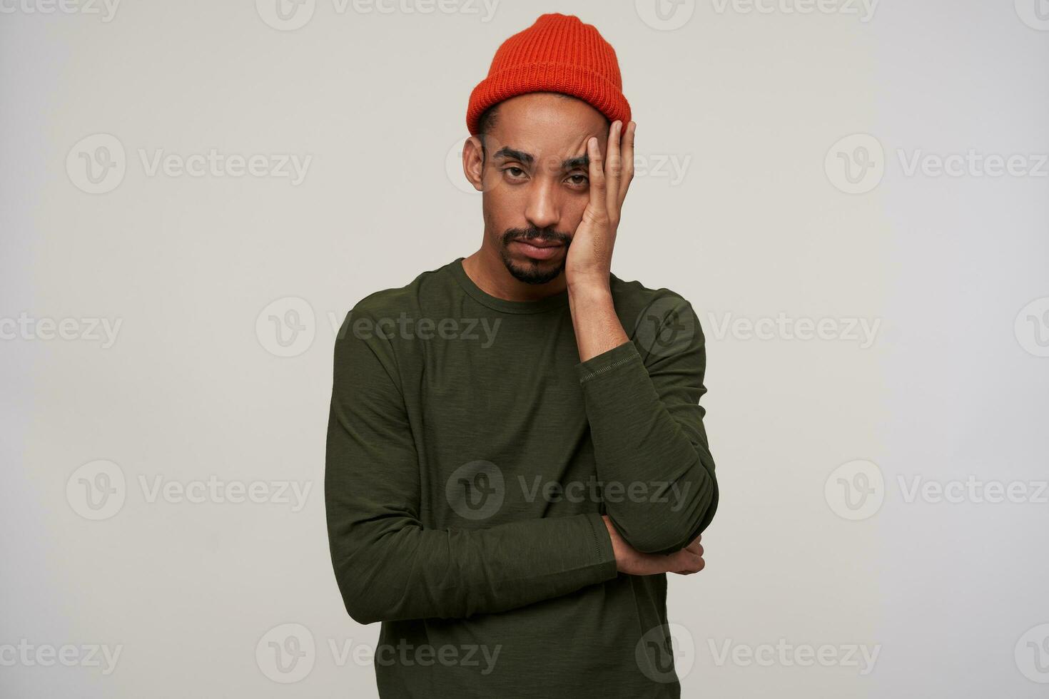 Exhausted young bearded dark skinned brunette male holding palm on his face and looking tiredly at camera, wearing casual clothes while posing over white background photo