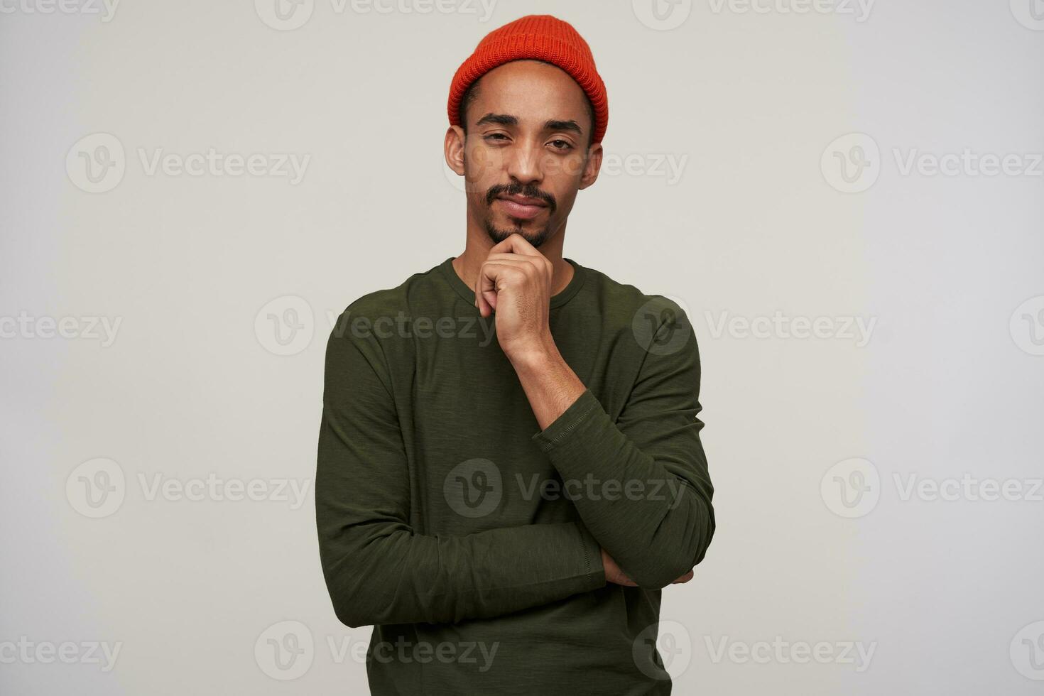Thoughtful young bearded dark skinned brunette guy holding his chin and squinting while looking at camera, wearing casual clothes while posing over white background photo