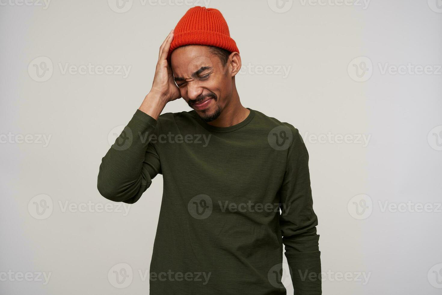 Excruciating young bearded handsome brunette man with dark skin keeping his eyes closed while suffering from heavy headache, holding palm on his head while posing over white background photo