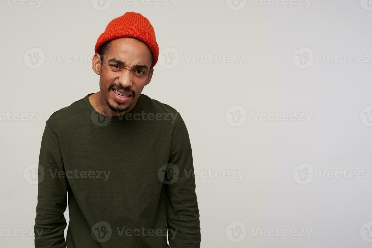 Indoor shot of young dark bearded dark skinned brunette guy looking to camera with dissatisfied gromace and frownig eyebrows, dressed in casual wear while standing over white background photo