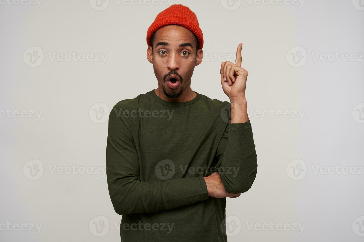 Indoor photo of excited young dark haired bearded man with dark skin raising up his forefinger and rounding his mouth while looking to camera, isolated over white background