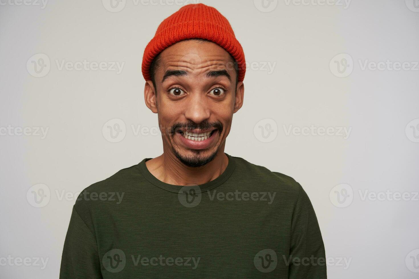 Open-eyed young pretty dark haired dark skinned male with beard wrinkling forehead with raised eyebrows while looking amazedly at camera, standing against white background photo