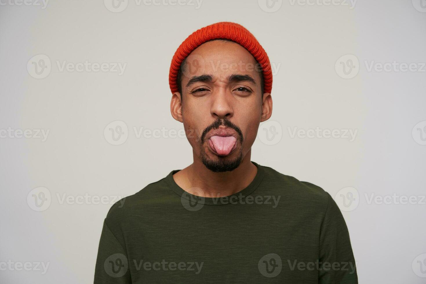 Portrait of young handsome dark skinned brunette guy in red hat with sticking out his tongue while looking to camera, isolated over white background in casual clothes photo