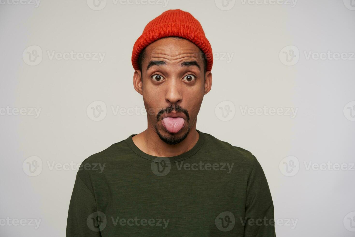 Close-up of young attractive bearded dark skinned man raising surprisedly eyebrows and showing his tongue to camera, making fun while posing over white background photo