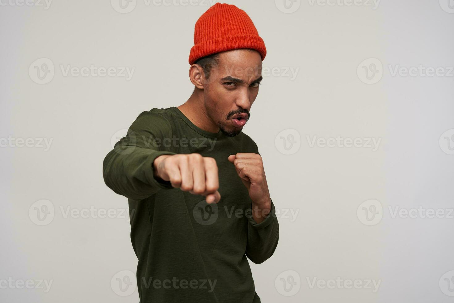 Young excited dark haired bearded male with dark skin boxing with raised fists and looking menacingly at camera, wearing casual clothes while standing over white background photo