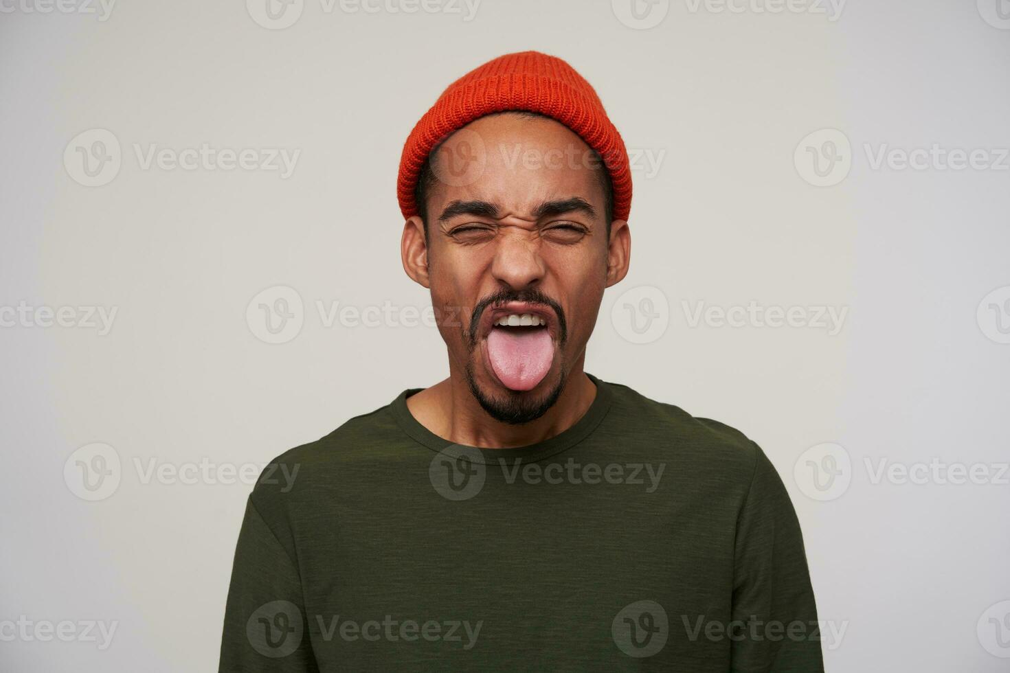 Indoor shot of young pretty bearded dark skinned guy frowning his face and showing tongue while expressing disgust, standing against white background in casual wear photo
