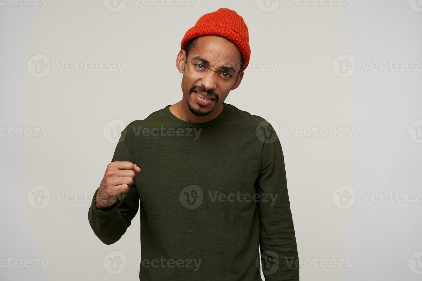 Portrait of young brown-eyed dark skinned brunette male with beard folding raised hand in fist and frowning his face while looking to camera, isolated over white background photo