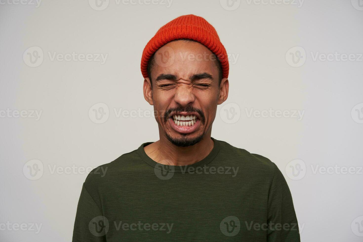 Studio photo of young dark haired bearded brunette man with dark skin showing disgust on his face while standing over white background in red hat and khaki pullover
