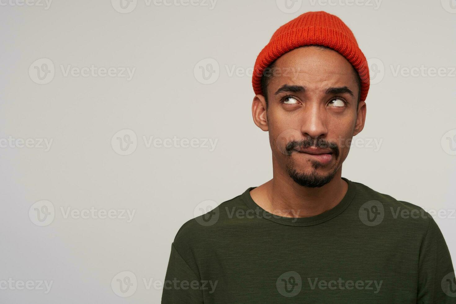 Puzzled young pretty dark skinned bearded brunette man biting underlip while looking upwards, plotting something, standing against white background in casual wear photo