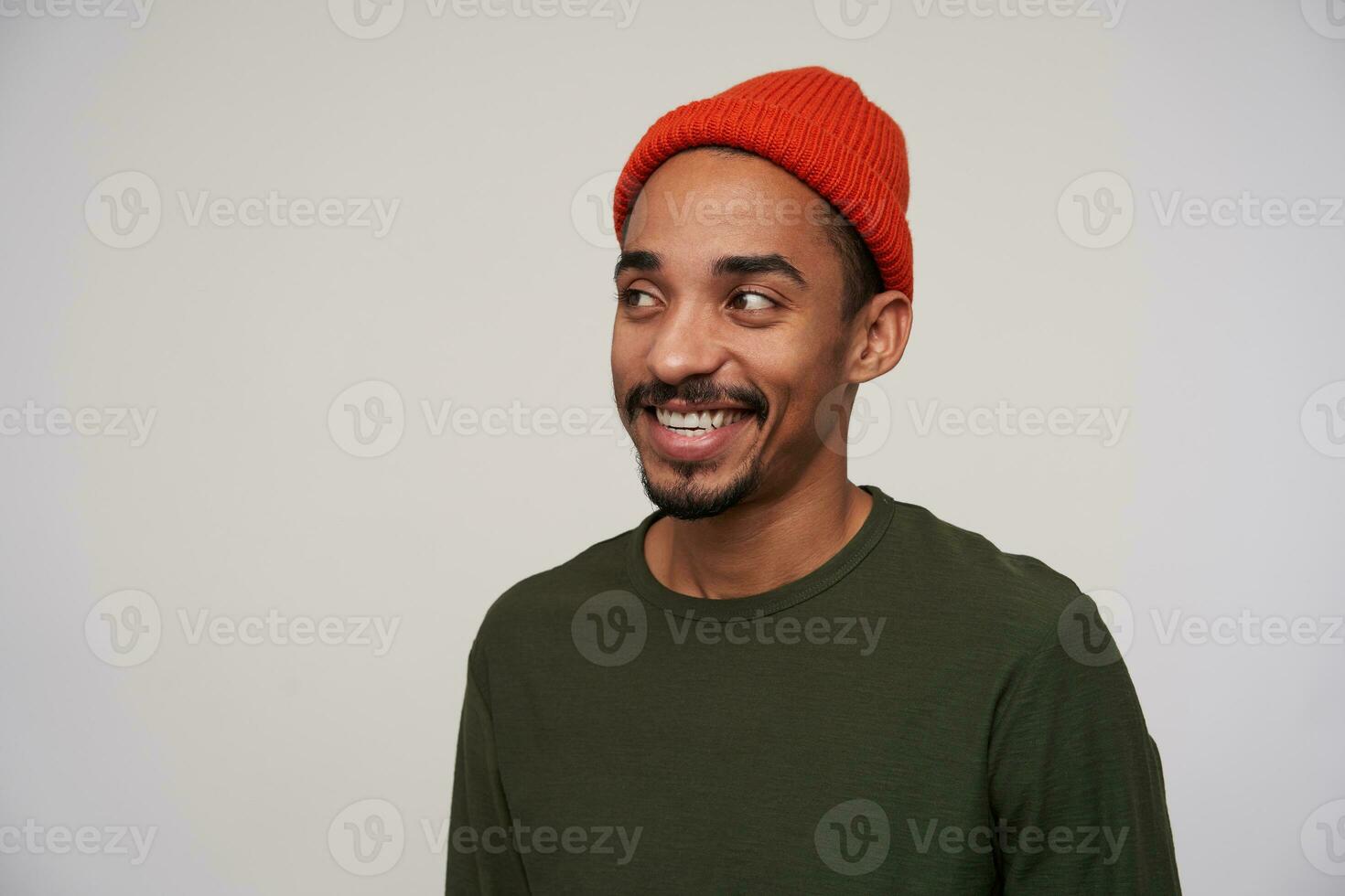 Close-up of charming young positive dark haired bearded man with dark skin looking happily aside with wide sincere smile, being in good mood while standing against white background photo