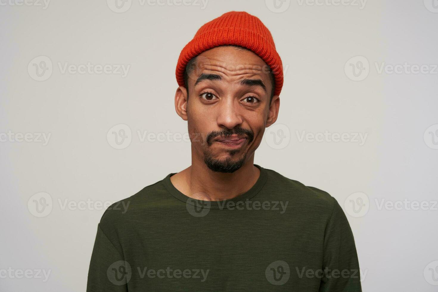 Portrait of young brown-eyed bearded dark skinned guy looking confusedly at camera and twisning his mouth, wrinkling forehead while posing over white background photo