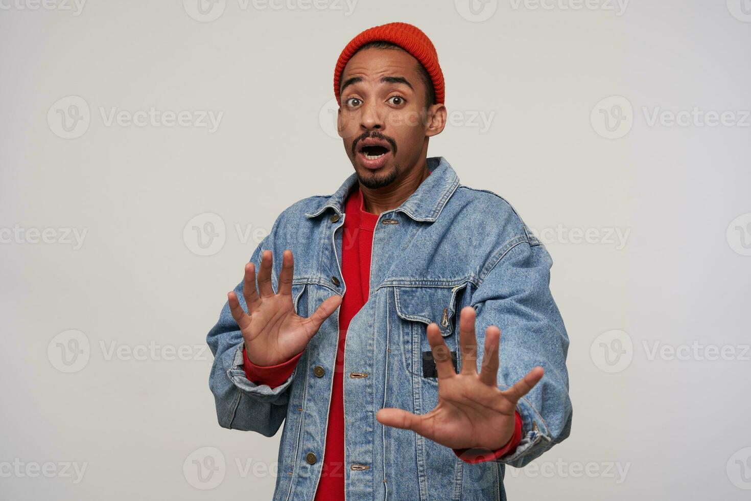 Fearful young brown-eyed dark skinned brunette man with beard protecting himself with raised hands and looking to camera with shocked face, isolated over white background photo