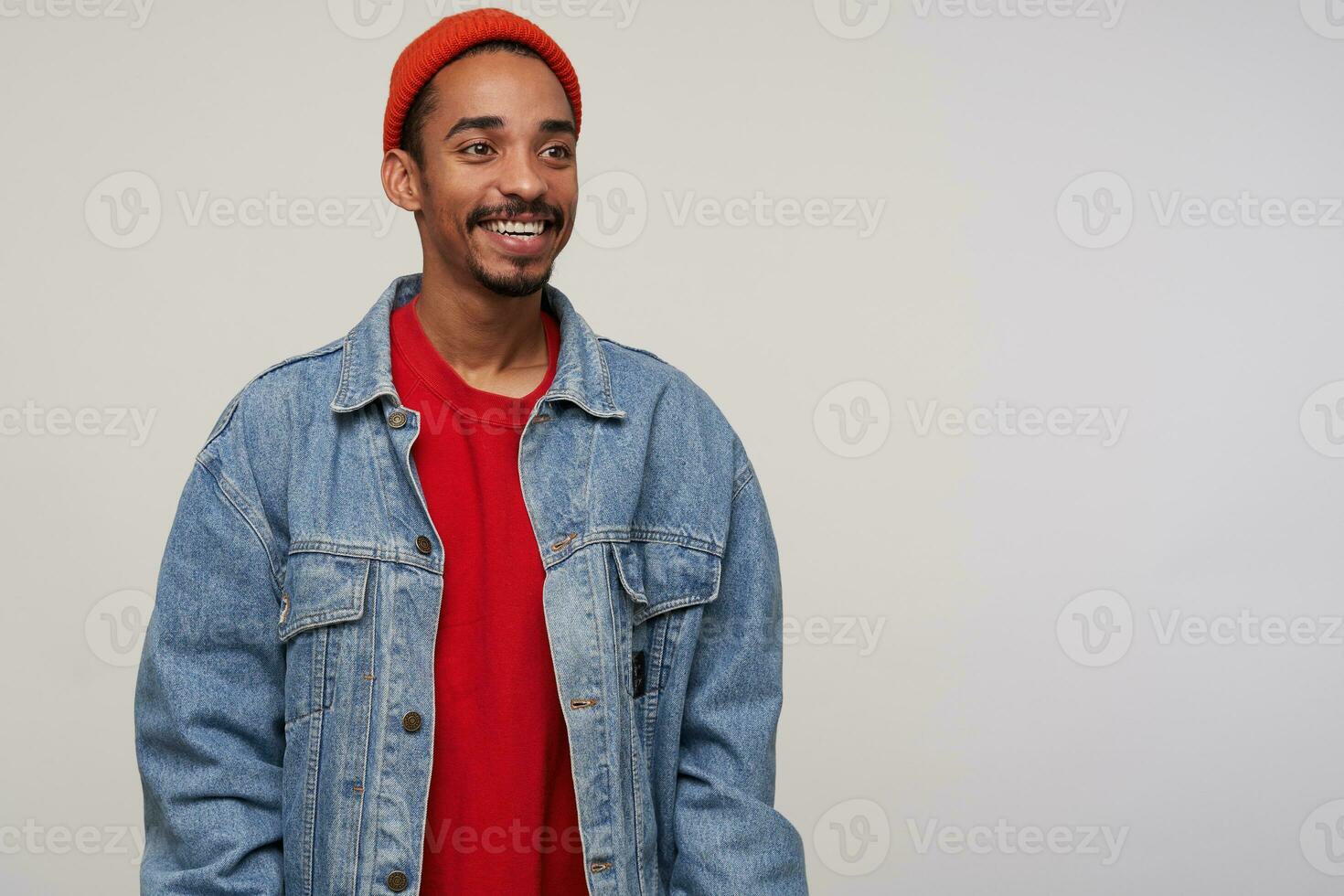 Cheerful attractive young dark haired bearded male with dark skin looking positivel aside with wide happy smile, dressed in red hat, red pullover and jeans coat while posing over white background photo